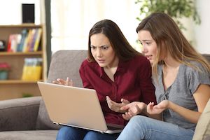 two friends sitting on couch looking at laptop confused
